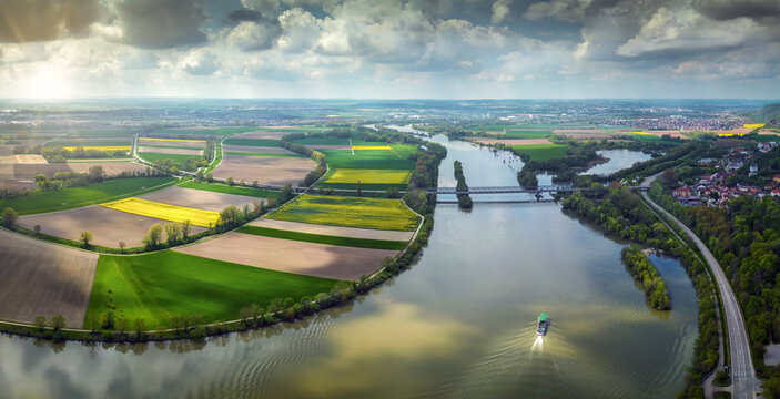 03 May 2023: View of a winding river flowing through vibrant green and yellow agricultural fields with a bridge crossing the water under a dramatic cloudy sky.