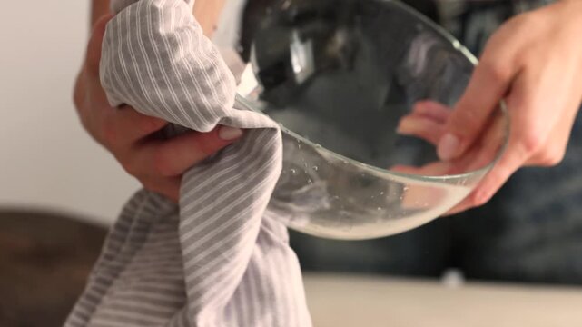 Woman wraps a striped cotton kitchen towel around a wet glass mixing bowl, wiping and drying it after washing, capturing simple everyday cleanup routine in a bright home kitchen setting.