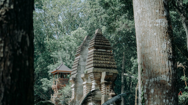 A picturesque view of the Candi Bentar split gate and a traditional Balinese Meru tower, partially obscured by a lush forest canopy and framed by large tree trunks in the foreground.