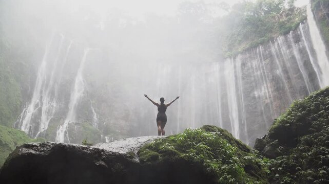 Young woman standing on a rock with arms outstretched, celebrating freedom at a powerful waterfall