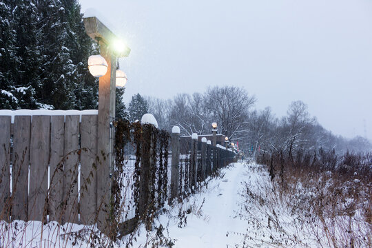 Glowing street lamps mounted on rustic wooden fence posts illuminate a path during a snowy winter evening, casting a warm light against the chilly, frost-covered forest backdrop