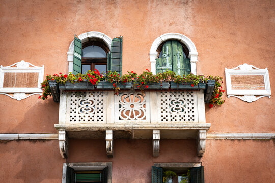 Europe - 04 October 2022: View of an ornate stone balcony with red geranium flowers and green shutters on a terracotta wall.
