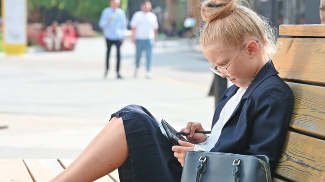A little girl dressed as a stylish businesswoman sits on a wooden street bench and writes something with a stylus on a digital tablet
