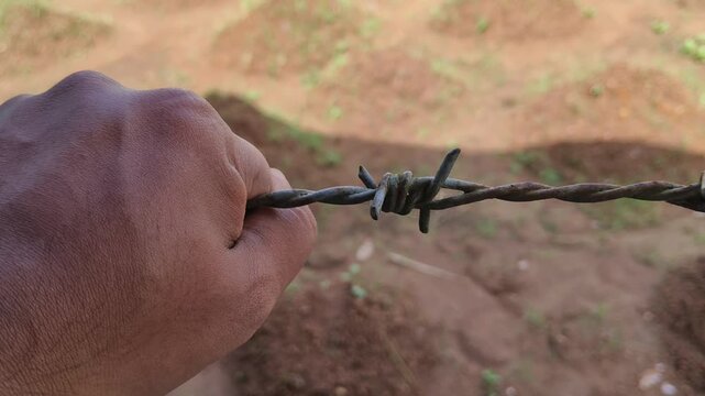 Poor native person evicted from tribal ancestral land territory after legal ownership conflict. Closeup of his hand holding tightly on new barbwire fence in his land. Save indigenous land concept.