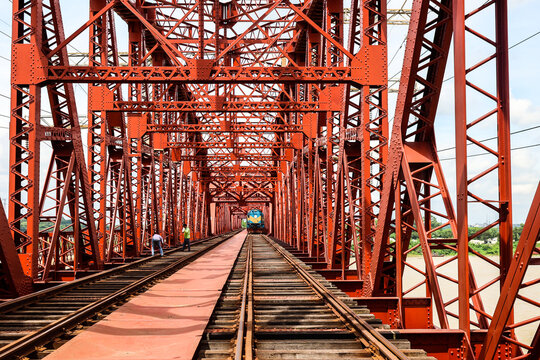 Pabna, Rajshahi Division, Bangladesh - 17 August 2025: View of the iconic red steel structure of the Hardinge Bridge with a train approaching in the distance and workers walking along the tracks.
