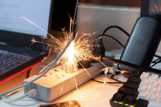Close-up of an electrical spark burning on overloaded power strip with messy cables on office desk, concept of short circuit fire hazard and workplace safety risk.