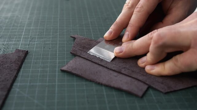 Professional leatherworker skiving genuine leather edges with a special knife on a cutting mat in a workshop