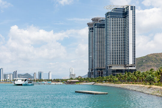Modern high-rise buildings rise along a waterfront, featuring a boat dock and lush tropical palm trees. The scene captures a vibrant coastal city with clear blue skies and distant mountains