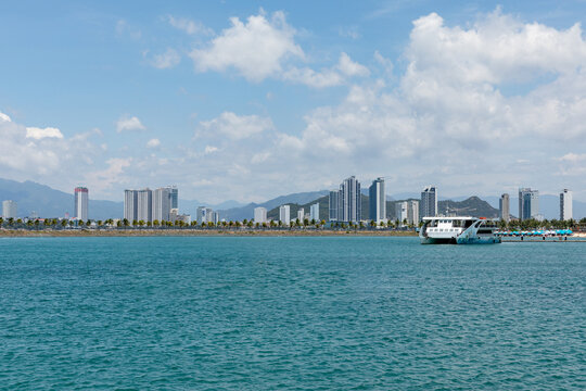 Small boat in marina, surrounded by a modern city skyline and mountains in the background, under a bright blue sky with fluffy clouds