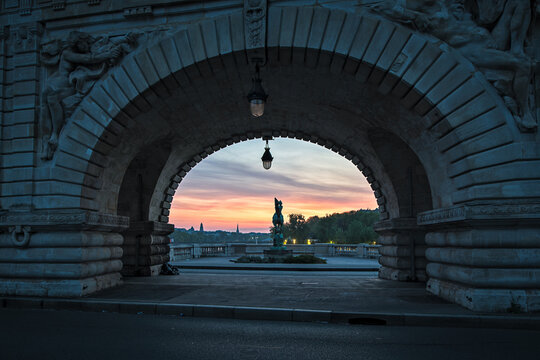 Paris, France - 30 April 2023: View of the Pont Alexandre III archway framing a bronze equestrian statue against a vibrant sunset sky over the Seine river.