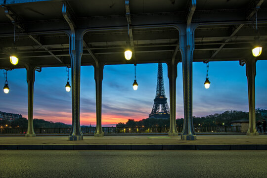 Paris, France - 30 April 2023: View of the Eiffel Tower framed by the iron structure of the Bir-Hakeim bridge at dusk with glowing street lamps and a colorful sunset sky.