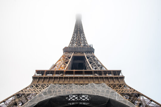 Paris, France - 29 April 2023: View of the Eiffel Tower with its intricate iron lattice structure partially obscured by low-hanging clouds and protective netting at the base.