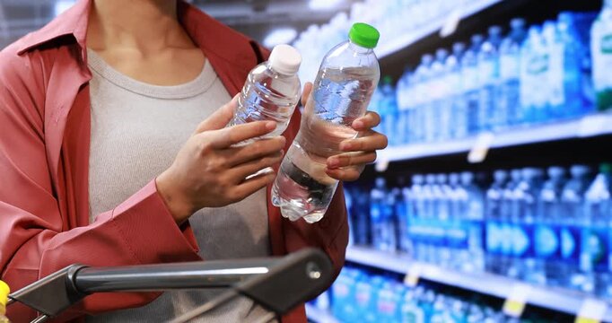 Person comparing two bottled water options while shopping in a supermarket aisle, with shelves of drinks and a shopping cart in the foreground