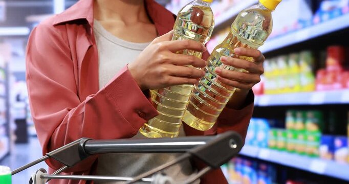 Person in a supermarket holding two bottles of cooking oil beside a shopping cart, with blurred grocery shelves in the background