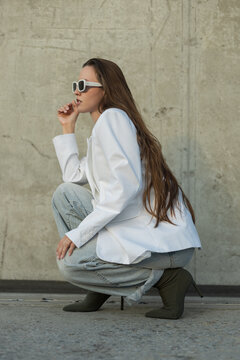 Stylish woman with long brown hair posing in side profile, wearing an oversized white blazer, light denim, high heels, and sunglasses against a concrete wall. Pretty fashion elegant girl