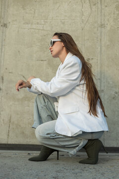 Side profile of a stylish young woman crouching outdoors against a concrete wall, wearing an oversized white blazer, light denim, high heels, and retro sunglasses.