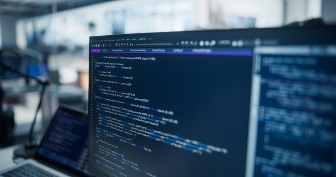 Close-up of Desktop Monitors Displaying Blue Sql and Php Source Code Beside a Laptop at a Software Engineer's Desk in a Diverse Open Plan Tech Office Where Developers Write, Test Debug Applications.