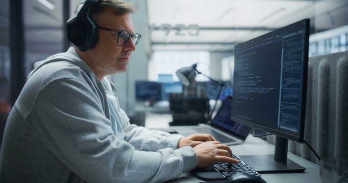 Portrait of a Middle-aged Caucasian Male Software Engineer in Hoodie, Glasses and Headphones Writing Code on a Desktop Computer, Developing Software Applications in a Diverse Office of Programmers.