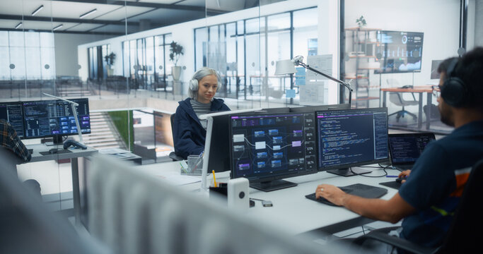 Caucasian Female Software Developer in Headphones Writes Code at Her Desk While an Indian Male Programmer Works on Dual Monitors Displaying Data Dashboards in a Diverse Office of Software Engineers.