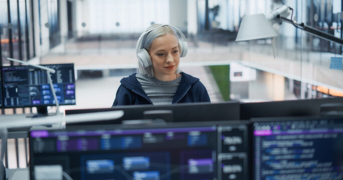 Portrait of a Young Caucasian Female Software Developer With Platinum Hair and Headphones Writing Code on Monitors Displaying Programming Scripts and Data Dashboards in Office of Software Engineers.