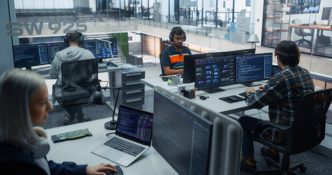 South Asian Male Software Engineer in Headphones Writes Code While a Senior White Female Developer and Caucasian Male Programmer Work on Data Dashboards and Applications in a Busy Tech Office.