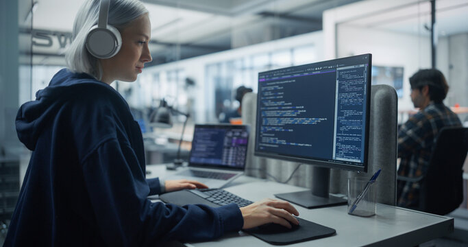 Portrait of a Young White Female Software Engineer With Silver Hair and Headphones Writing Program Code on a Desktop Computer and Laptop, Developing a Web Application in an Office With Colleagues.