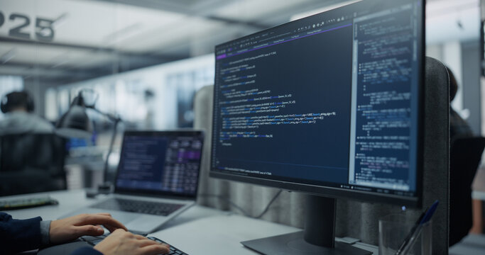 Close-up of a Desktop Computer Screen Displaying Program Code and Database Script While the Hands of an Unrecognizable Software Engineer Type on a Laptop in an Office With Diverse Programmers Working.