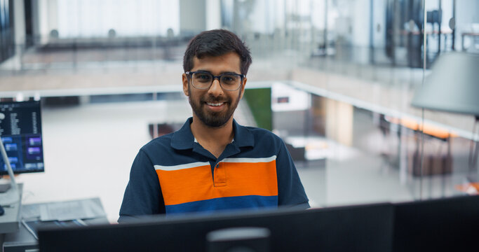 Friendly Indian Male Software Developer in Glasses Looking at Camera With a Warm Smile While Writing Program Code on Desktop Computers, Creating Digital Products With a Engineering Team in an Office.