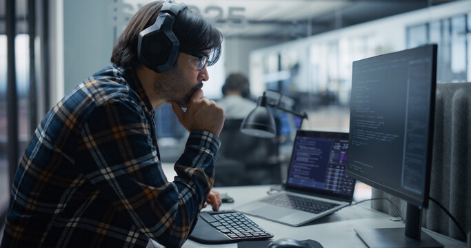 Thoughtful Young Indian Male Software Engineer in Headphones and Plaid Shirt Analyzes Program Code on Computer Screens, Creating Software Solutions and Working With a Team of Developers in an Office.