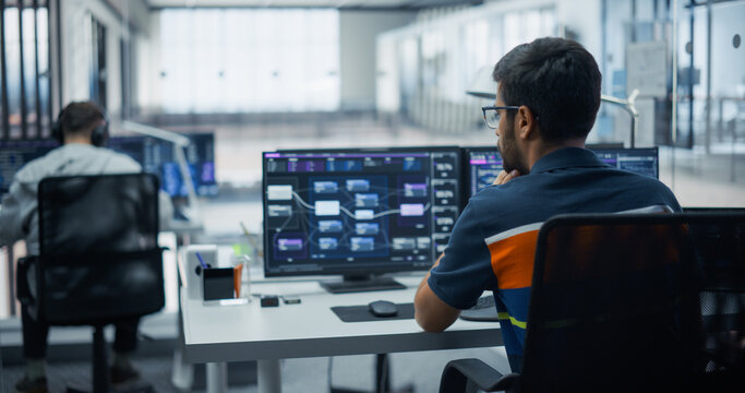 Pensive Indian Male Software Engineer in Glasses Reviews Application Architecture Diagrams on Dual Computer Screens, Writing Program Code and Building Software With a Team of Developers in an Office.