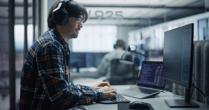 Focused Indian Male Software Developer in Headphones and Glasses Writes Program Code on Computer Screens, Building Applications and Solving Technical Tasks Alongside a Team of Engineers in an Office.