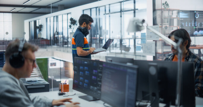 Multiethnic Group of Software Developers in Office: Indian Male Engineer Stands Reviewing Code on Laptop, Caucasian Male Coder With Headphones Types Programming Language on Desktop Computer Monitors.