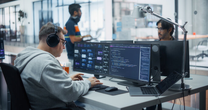 Male Software Engineer in Hoodie and Headphones Listens to Music While Typing Program Code on Dual Monitors and Laptop, Working on Data Analytics System in Office With Multicultural Colleagues.