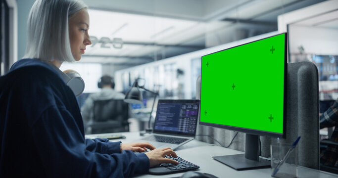 Portrait of White Female Software Developer With Headphones Writing Program Code on Green Screen Chroma Key Computer and Laptop at Her Desk in an Office With Team of Software Engineers.