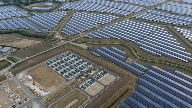 Aerial view of a large solar farm featuring extensive rows of photovoltaic panels, battery storage containers, and electrical substation infrastructure in Faversham, England, United Kingdom.
