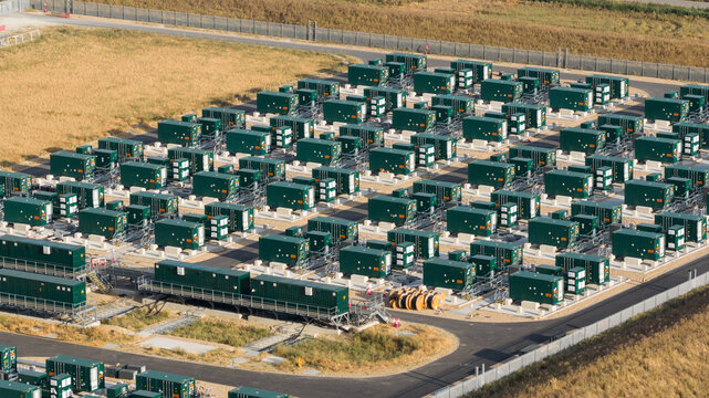 Aerial view of a large battery energy storage system facility with rows of green containerized battery units on a gravel site in Faversham, England, United Kingdom.