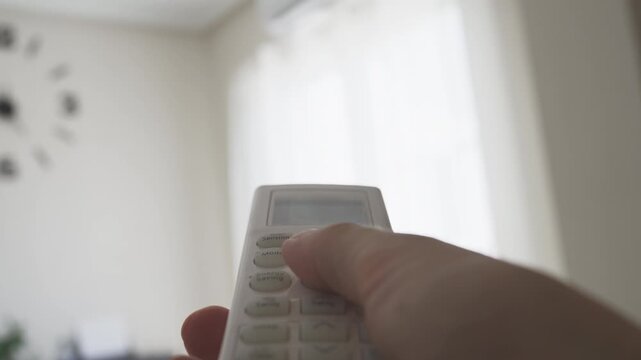 Close-up of a person's hand holding a remote control and pressing the red power button to activate the air conditioner, adjusting the home climate for better comfort and temperature