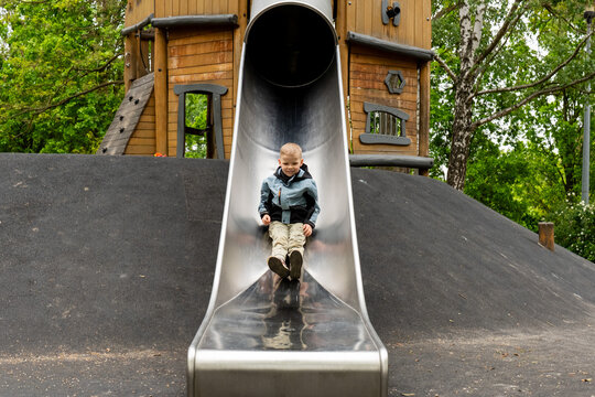 Happy young boy sliding down large metal slide at outdoor playground surrounded by green trees. Concept of active childhood, outdoor recreation, physical activity, fun and healthy lifestyle. 