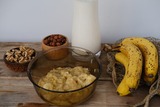 Slicing ripe bananas on wooden cutting board for healthy breakfast with fresh natural ingredients, glass milk and bowl of hazelnuts on rustic table, cooking process and nutritious morning meal concept