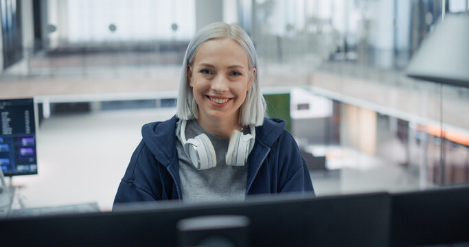 Portrait of a Cheerful Caucasian Female Software Engineer With Platinum Hair and White Headphones Around Her Neck Smiling at Camera While Writing Program Code on a Desktop Computer in a Office.