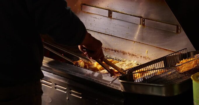Close up rear view shot of street food vendor moving large breaded meat cutlets around inside commercial deep fryer. Cook uses long metal tongs to separate and distribute frying pieces in bubbling oil