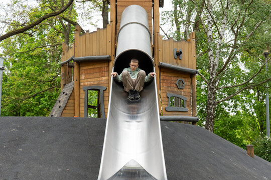 School age boy with glasses sliding down large metal tube slide from wooden adventure tower in green park. Concept of active childhood, courage, physical development and outdoor recreation.