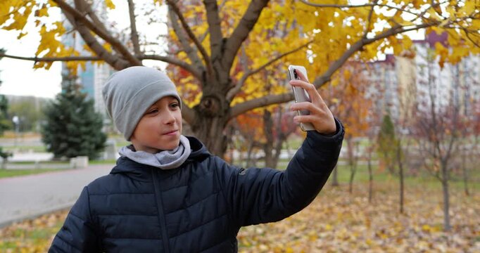 Smiling schoolboy takes selfie in autumn park, enjoying colorful fall leaves and outdoor leisure. Casual backpack and seasonal setting create relatable school life and youth lifestyle. Happy child.