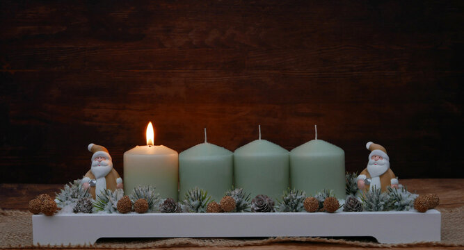 1st Sunday of Advent: Advent wreath with four green candles, the first one lit featuring pine cones, small fir sprigs, and two Santa Claus figures, set against a wooden background with space for text.