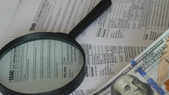 Close-up shot of a magnifying glass and a stack of hundred-dollar bills lying on US Individual Income Tax Return Form 1040 forms. 