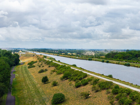Aerial view of Mittellandkanal fork in Magdeburg with calm waterway, embankments, paths and dry summer grass. Broad canal landscape stretches beneath layered clouds, greenery and distant ships