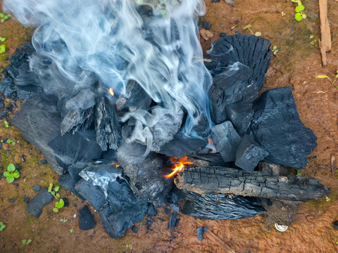 Smoldering charcoal and burning wood produce white smoke on damp brown soil, creating rustic outdoor fire scene with glowing ember and charred texture