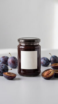 Glass jar of homemade plum jam with blank white label surrounded by fresh ripe plums on grey background