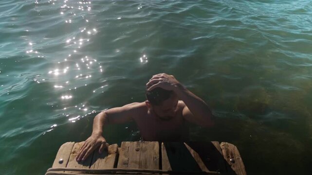 Man finishing a swim holds onto a wooden pier near the shore as boats rock gently on the waves, creating a peaceful seaside and summer vacation atmosphere.