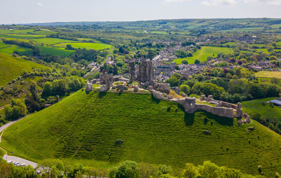 Aerial view of Corfe Castle ruins perched on a grassy hill overlooking the village and the lush green Purbeck countryside under a bright sky, Corfe Castle, England, United Kingdom.